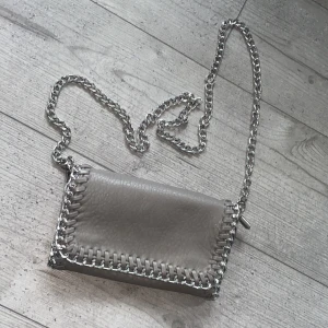 Grå axelväska med kedjedetaljer - Snygg grå axelväska från Tiamo i skinnimitation med silverfärgad kedja som axelrem och runt kanterna. Väskan har magnetlås, innerfack med dragkedja och plats för mobil och småprylar. Perfekt storlek för det viktigaste och ger en cool vibe till din outfit.