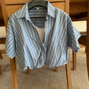 Blåvit randig skjorta från Twist - Säljer en blå skjorta med vita ränder från Twist. Skjortan har korta ärmar, klassisk krage och knäppning framtill. Modellen är croppad och passar perfekt till sommaren. Superfräsch och enkel att styla med jeans eller kjol.