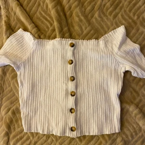 Vit ribbad croppad topp med knappar - Säljer en vit croppad topp med ribbad struktur och bruna knappar framtill. Toppen har korta ärmar och en offshoulder-design som ger en trendig look. Perfekt till sommaren!