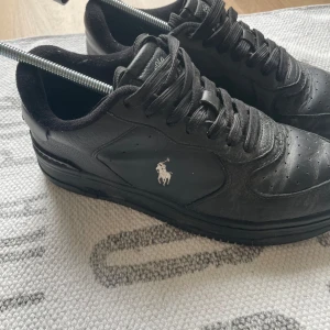 Svarta sneakers från Polo Ralph Lauren - Stilrena svarta sneakers från Polo Ralph Lauren i skinn med klassisk siluett. Skorna har svart snörning, perforerad tå och Polo-logga i vitt på sidan samt broderad Polo-text på hälen. Perfekta för en clean och sportig look.