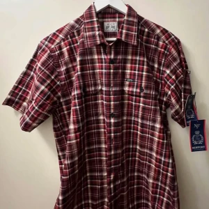 Rutig kortärmad skjorta - Snygg röd och vit rutig skjorta med korta ärmar och klassisk krage. Skjortan har knappar framtill och två bröstfickor med lock. Perfekt för dig som gillar en avslappnad men ändå stilren look.