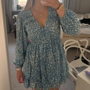 Blå Zara klänning  - Supersöt blå klänning med vitt leopardliknande mönster från Zara. Klänningen har lång ärm, v-ringning och volangdetaljer nertill. Den har en smickrande passform med markerad midja och lätt utställd kjol. Perfekt för dig som gillar färg och mönster!