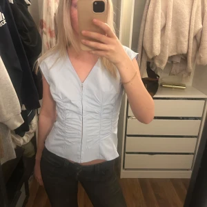 Ljusblå v-ringad blus - Säljer en ljusblå blus med v-ringning och korta ärmar. Toppen har figurnära passform och fina rynkade detaljer framtill. Perfekt för en stilren och fräsch look.