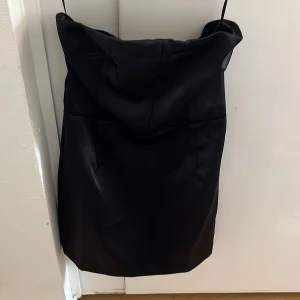 Svart axelbandslös kortklänning - Säljer en svart axelbandslös kortklänning med enkel och stilren design. Klänningen har smala axelband och en rak passform som ger en snygg siluett. Perfekt för dig som gillar minimalistisk stil.