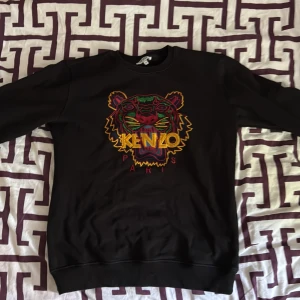 Svart Kenzo sweatshirt med tigerbrodyr - Svart sweatshirt från Kenzo med ikonisk broderad tiger i färgerna gul, röd, grön och blå på bröstet. Klassisk rund hals och långa ärmar. Perfekt statement-plagg för dig som vill sticka ut med en fet logga och färgglad design.