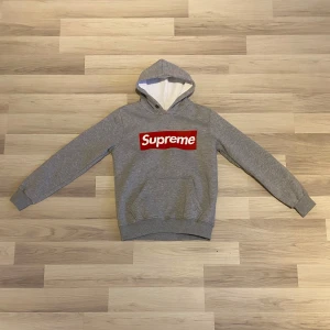 Grå hoodie från Supreme - Säljer en grå hoodie från Supreme med den klassiska röda boxloggan framtill. Storleken är L i ungdoms kriterier. Tröjan har huva och långa ärmar, samt ribbade muddar vid ärmslut och nederkant. Perfekt för en streetwear-look.