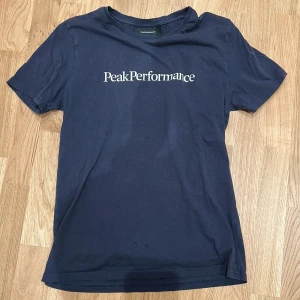Mörkblå t-shirt från Peak Performance - Säljer en mörkblå t-shirt från Peak Performance med vit logotyp på bröstet. T-shirten har klassisk passform och rund hals. Perfekt för en sportig och avslappnad stil. Obs små hål längst ned 