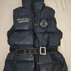 Mörkblå dunväst från HV Polo med bälte - Snygg mörkblå dunväst från HV Polo med broderade loggor fram och bak. Västen har hög krage, dragkedja och knappar samt ett brett bälte i midjan för extra stil. Perfekt för lager-på-lager och en sportig look.