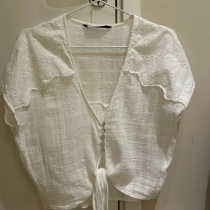 Vit blus med broderad krage och knyt - Säljer en söt vit blus med stor broderad krage och knyt framtill. Blusen har korta ärmar, dekorativa knappar och romantiska broderidetaljer på kragen. Perfekt för dig som gillar en feminin och vintageinspirerad stil.