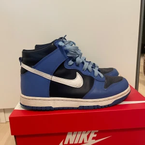 Nike Dunk High GS blå/vita sneakers - Nike Dunk High GS i medium blue och vitt med marinblå detaljer. Klassisk high-top siluett med vit swoosh, blå snörning och slitstark ovandel i skinn. Perfekta för dig som gillar streetstyle och vill sticka ut med snygga färgblock. Bra skick endast använda fåtal gånger. Säljer fö de blivit för små. Pris kan diskuteras 