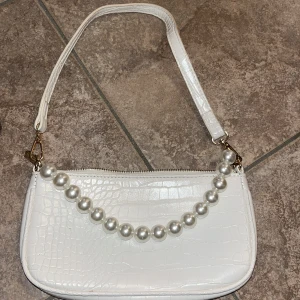 Vit axelväska med pärldetalj - Snygg vit axelväska i croco-mönstrat skinnimitation med guldfärgade detaljer och en avtagbar rem med stora pärlor. Väskan har dragkedja upptill och en stilren, rektangulär form som ger en trendig vibe.