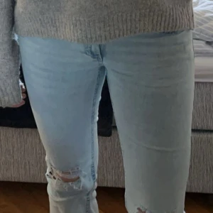Ljusblå bootcut jeans med slitningar - Säljer ett par ljusblå bootcut jeans med slitningar på knäna och broderade detaljer på bakfickorna. Jeansen har låg midja och klassisk femficksdesign. Perfekta för dig som gillar en avslappnad och trendig stil.