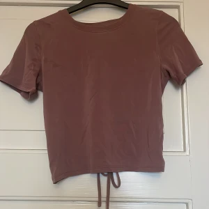 Lila croppad topp med knyt från Lululemon - Säljer en lila croppad topp från Lululemon med korta ärmar och rund halsringning. Toppen har en snygg öppen rygg med knytband nedtill, perfekt för en trendig look.