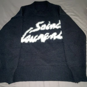 Mörkgrå stickad tröja från Saint Laurent - Säljer en mörkgrå stickad tröja från Saint Laurent med vit logotyp framtill i ett coolt, suddigt mönster. Tröjan har rund halsringning och breda ribbade muddar vid ärmslut och nederkant. Perfekt för dig som gillar exklusiva och trendiga plagg.