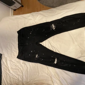 Svarta ripped skinny jeans med färgstänk - Svarta skinny jeans med slitna detaljer och hål på benen, samt vita färgstänk för en edgy look. Klassisk femficksmodell med dragkedja och knapp. Perfekta för dig som gillar streetstyle och vill sticka ut.