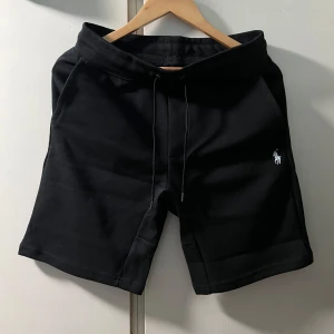 Svarta shorts från Polo Ralph Lauren - Svarta shorts från Polo Ralph Lauren med slim fit och klassisk broderad logga i vitt på ena benet. De har snörning i midjan och är tillverkade i mjukt material, perfekta för en chill och stilren look. Bakficka och två sidofickor för extra funktion.