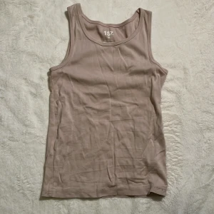 Beige linnetopp från 157, stl 150 - En basic beige linnetopp från 157 i 100% ekologisk bomull. Modellen är ärmlös med klassisk rund halsringning och mjuk känsla. Perfekt att styla med jeans eller shorts för en chill look.