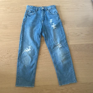Blå slitna jeans från Zara Denim - Säljer ett par blå jeans från Zara  med raka ben och råa kanter nertill. Jeansen har slitningar och hål på framsidan för en cool look. Klassisk femficksmodell och knappgylf.Byxorna är väldigt sköna men ändå snygga