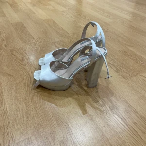 Vita klackskor med öppen tå - Säljer ett par vita klackskor med hög blockklack och öppen tå. Skorna har en elegant rem runt vristen och är tillverkade i ett glansigt material som ger en lyxig känsla. Perfekta för dig som vill sticka ut med stilrena detaljer och modern design.