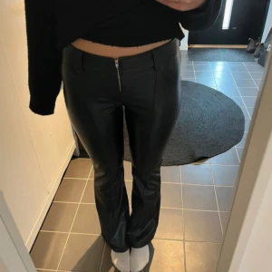 Svarta bootcut skinnbyxor  - Snygga svarta lågmidjade byxor i skinnimitation med bootcut passform och dragkedja framtill. Byxorna har en tight passning upptill och släpper ut vid benen för en trendig siluett. Perfekta för dig som vill ha en edgy look.