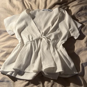 Vit omlottopp med knyt och volang - Supersöt vit omlottopp med knyt i midjan och volangig nederdel. Toppen har korta ärmar och är gjord i ett ribbat bomullstyg som känns både luftigt och mjukt. Perfekt för dig som gillar en feminin och avslappnad stil.💗💖