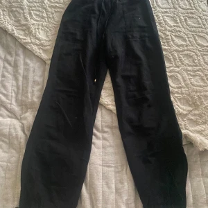 Svarta vida linnebyxor med snörning - Svarta linnebyxor med avslappnad passform och breda ben. Byxorna har resår i midjan och snörning för extra komfort. Perfekta för en chill och stilren look, lätta att matcha med sneakers eller sandaler. Snör grejen vid fotleden åt vänster är lite kortare.