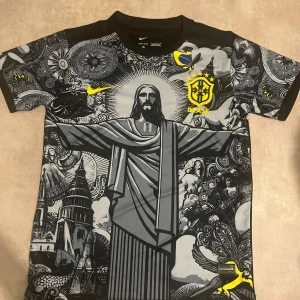 Svart Brasilien fotbollströja  - Unik svart kortärmad Brasilien fotbollströja från Nike med stort tryck av Kristusstatyn och detaljerade grafiska mönster i grått och vitt. Gula Brasil-logga och Nike-märke på bröstet. Tillverkad i Dri-Fit material för bra komfort.