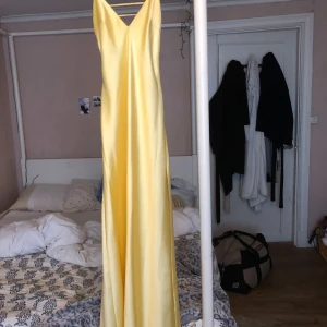 Lång gul satin balklänning  - Snygg långklänning i ljusgul satin med smala axelband och v-ringning både fram och bak. Klänningen har en elegant, slät yta och faller mjukt längs kroppen. Perfekt för dig som vill sticka ut med en fräsch och somrig look.Använd bara en gång och är i ny skick 