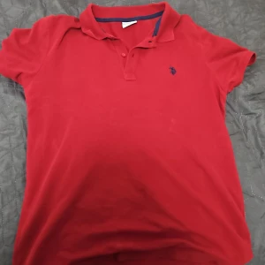 Röd pikétröja från U.S. Polo Assn - Säljer en klassisk röd pikétröja från U.S. Polo Assn i storlek M. Tröjan har korta ärmar, krage med knappar och en liten blå logga broderad på bröstet. Perfekt för en sportig och stilren look.