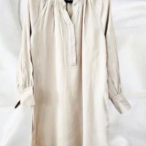 Ljusbeige långärmad skjortklänning - Luftig och stilren skjortklänning i ljusbeige från ARKET. Klänningen har långa ärmar med manschett, knappar framtill och fina rynkade detaljer vid axlar och halsringning. Tillverkad i mjukt, glansigt material som ger en lyxig känsla.