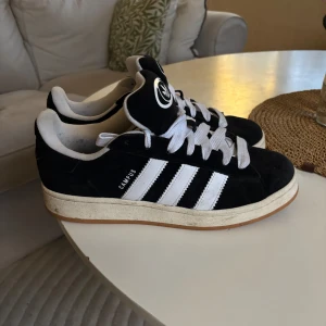 Adidas Campus sneakers svart/vit strl 44 - Använda max 5 gånger då jag fick ett par lika dana i present. Svarta Adidas Campus sneakers i mocka med klassiska vita ränder och vit logga på plösen. Skorna har vita snören och en brun gummisula. Perfekt för dig som gillar retrostil och streetwear. Storlek 44 (EU). Jag står för frakten 