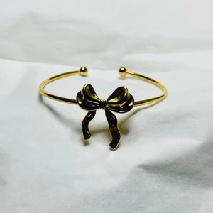 Guldfärgat armband med rosett - Snyggt armband i guldfärg med en dekorativ rosett framtill. Armbandet är öppet och har små kulor i ändarna för en stilren look. Perfekt accessoar för att lyfta din outfit.