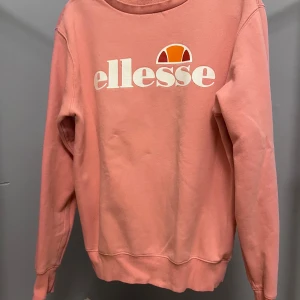 Rosa sweatshirt från Ellesse - Säljer en rosa sweatshirt från Ellesse med stort vitt logotryck på bröstet och orange detalj. Tröjan har rund hals, långa ärmar och är gjord i mjuk bomullsblandning med borstad insida för extra komfort. Perfekt för en chill och sportig look.