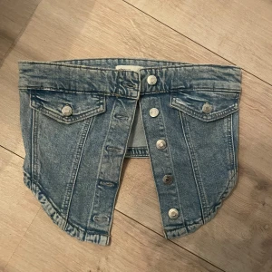 Kort jeanstopp med knappar - PRIS GÅR ATT DISKUTERA. Cool kort jeanstopp i blå denim med silverfärgade knappar framtill. Toppen har två fickor med lock och knapp på framsidan för en extra edgy look. Perfekt för dig som vill sticka ut med en unik stil.