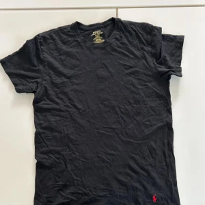 Svart t-shirt från Polo Ralph Lauren - Svart t-shirt från Polo Ralph Lauren med klassisk passform och rund hals. Tillverkad i 100% bomull och har den ikoniska röda Polo-loggan broderad längst ner. Perfekt basic med stilren look.