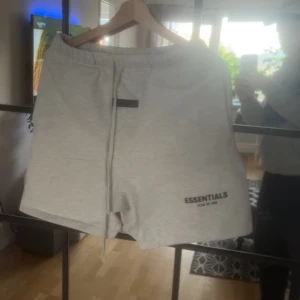 Grå shorts från Essentials Fear of God - Snygga ljusgrå shorts från Essentials Fear of God med tryckt logga på ena benet och långa snören i midjan. Modellen har en normal passform och är tillverkad i mjukt bomullsmaterial. Perfekta för en avslappnad stil.