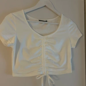 Vit croppad topp med snörning - Superfin vit croppad topp med korta ärmar och snygg snörning framtill som ger en rynkad effekt. Djup rundringning och knyt längst ner. Perfekt till jeans eller kjol för en fräsch look.