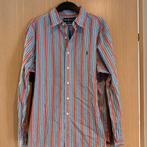 Randig skjorta från Ralph Lauren XL - Färgglad skjorta från Ralph Lauren i klassisk passform. Skjortan har vertikala ränder i blått, rött, grönt, vitt och orange samt knappar framtill. Tillverkad i bomull och med lång ärm. Perfekt för dig som gillar en stilren men ändå lekfull look.