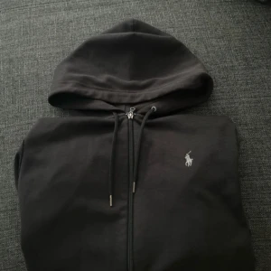 Svart hoodie från Ralph Lauren - Svart hoodie från Ralph Lauren med dragkedja och huva. Klassisk design med vit broderad logga på bröstet och snörning i huvan. Perfekt för en chill och stilren look. Tillverkad i mjuk bomull för extra komfort.