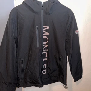 Svart vindjacka från Moncler - Svart vindjacka från Moncler med stor logga längs framsidan. Jackan har huva, tre dragkedjefickor och justerbar nederkant. Klassisk Moncler-patch på ärmen och snygg, sportig look. Perfekt för dig som gillar streetwear och exklusiva märken.