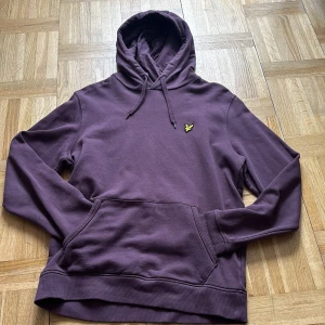 Lila hoodie från Lyle & Scott - Säljer en snygg lila hoodie från Lyle & Scott med klassisk gul logga på bröstet. Tröjan har huva med snörning, känguruficka och är tillverkad i mjuk bomull. Perfekt för dig som gillar en chill och stilren look.