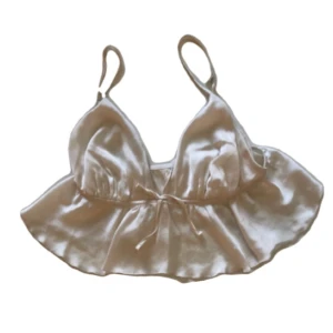 Vit satin linnetopp - Snygg linnetopp i glansig vit satin med smala axelband och volangkant nedtill. Toppen har en söt knytning framtill och en lätt croppad passform. Perfekt för dig som vill ha en trendig och feminin look.