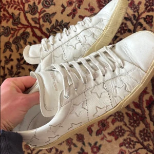 Saint Laurent Star - Snygga vita sneakers från Saint Laurent med stjärnmönster i skinn. Klassisk modell med snörning och platt sula. Perfekta för dig som gillar en clean look med en twist. Märkeslogga på plösen och sula i beige ton.