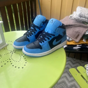Nike Air Jordan 1 Mid blå och svart - Säljer ett par Nike Air Jordan 1 Mid i blått och svart med vita detaljer. Skorna har klassisk snörning, svart swoosh och rund tå. Perfekta för dig som gillar streetwear och vill sticka ut med färg. Ikonisk siluett och bekväm passform.