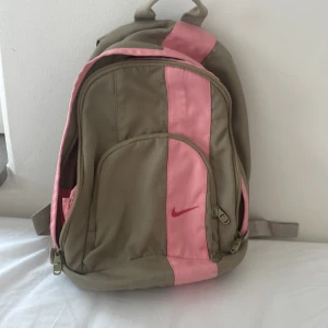 Nike ryggsäck i beige och rosa - Snygg ryggsäck från Nike i beige med rosa detaljer och röd logga framtill. Väskan har flera fack med dragkedja, vadderade axelband och rosa innerfoder med broderad Nike-logga. Perfekt för skolan eller träning, rymmer allt du behöver.