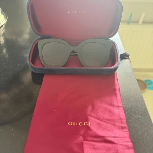 Svarta solglasögon från Gucci - Snygga svarta solglasögon från Gucci med fyrkantig form och breda skalmar. På sidorna finns guldfärgade GG-loggor och två silverfärgade stjärnor. Kommer med originalfodral i blå sammet och röd påse med guldtext. Perfekt accessoar för att lyfta din stil.
