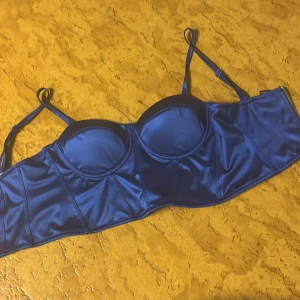 Blå satin korsettopp med axelband - Snygg blå korsettopp i glansigt satinmaterial med justerbara axelband och formade kupor. Toppen har en croppad passform och markerade sömmar som ger en cool siluett. Perfekt att styla med höga jeans eller kjol för en trendig look.