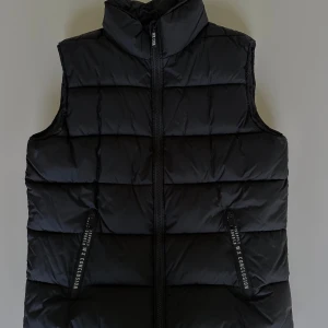Svart dunväst  - Jag säljer denna svarta dunväst med hög krage, normal passform och snygga textdetaljer vid fickorna. Perfekt att ha över en hoodie när det är lite varmare ute!☀️🌷