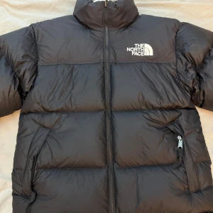 THE NORTH FACE 1996 RETRO NUPTSE JACKET - Säljer en svart dunjacka från The North Face med hög krage och dragkedja framtill. Jackan har två stora fickor med dragkedja och broderad logga på bröstet. Perfekt för kalla dagar och riktigt skön att bära.