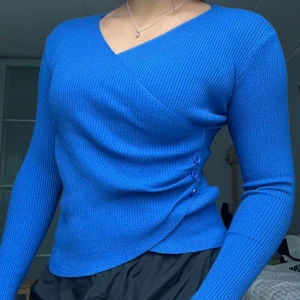 Blå omlottopp med knappar - Säljer en supervacker och SUPERSKÖN blå omlottopp med ribbad struktur och dekorativa knappar i sidan. Fick den i present men i för liten storlek. Den är helt oanvänd.